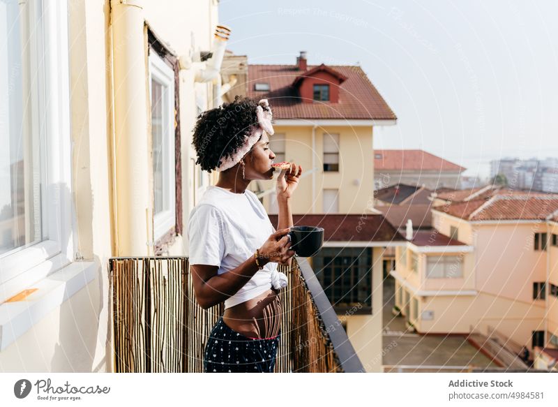 Black woman having breakfast on balcony drink toast apartment ethnic mug sip eat female slim rest relax lifestyle street joy weekend energy beverage lady