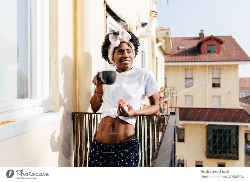 Black woman having breakfast on balcony drink toast apartment ethnic mug sip eat female slim rest relax lifestyle street joy weekend energy beverage lady