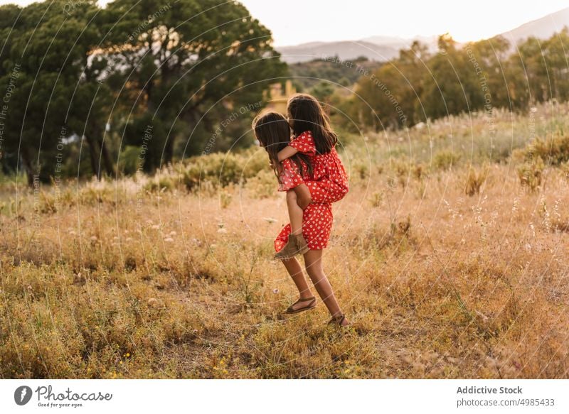 Smiling girl carrying sister on back in field piggyback summer nature ride vacation happy carefree friend fun meadow red walk family dress sibling together ears