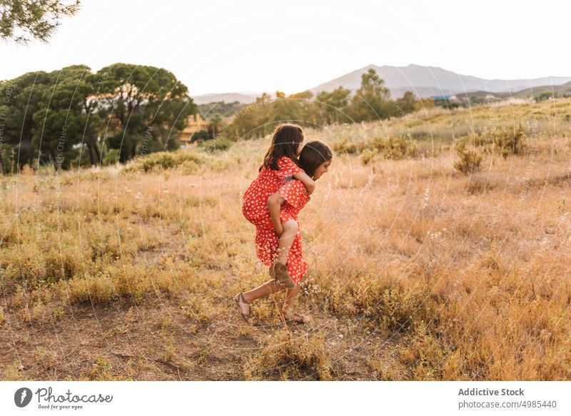 Smiling girl carrying sister on back in field piggyback summer nature ride vacation happy carefree friend fun meadow red walk family dress sibling together ears