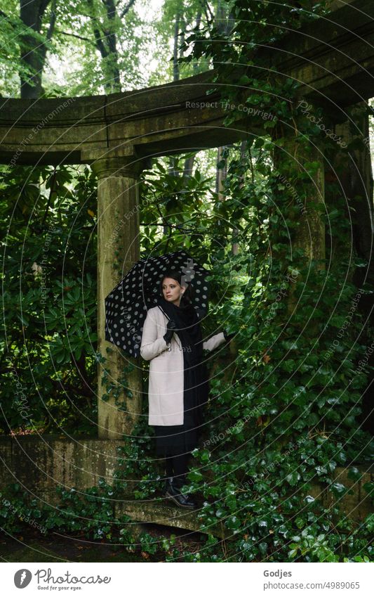 [HH Unnamed Road] Young woman with umbrella stands in arch of columns overgrown with ivy Woman Umbrella Stele Column Leaf canopy Monument Exterior shot Gray