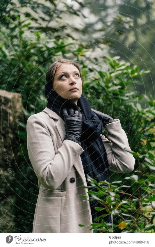 [HH Unnamed Road] Portrait of a young woman in a beige coat against a green background. She looks up and adjusts her scarf Woman person Coat Scarf look up