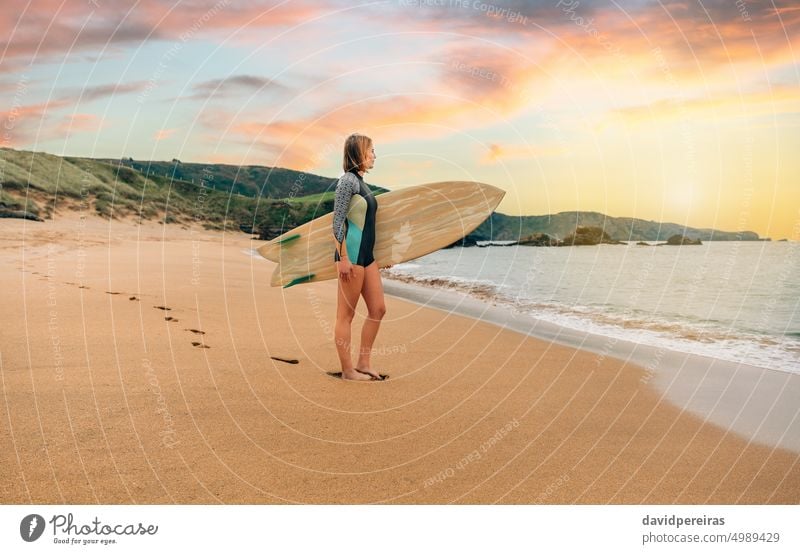 Surfer woman with wetsuit carrying surfboard looking to the sea at the beach young surfer back sand footprints posing people 20s outdoors sport surfing suit