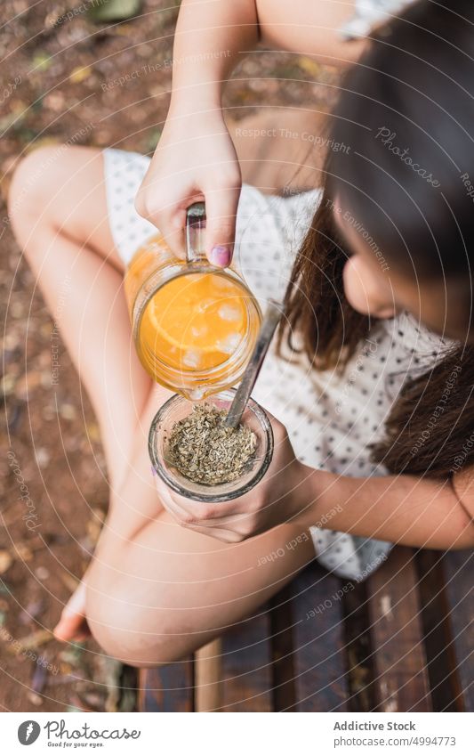 Crop girl pouring lemonade into mate on bench in daytime refreshment drink tea natural herbal ground leaf beverage legs crossed calabash gourd childhood kid jug