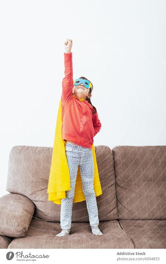 Girl in mask and cape pretending to be superhero girl costume confident childhood room kid sofa light home white wall little imagination couch courage fearless