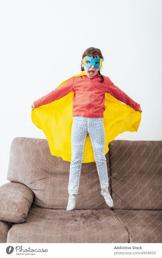 Girl in mask and cape pretending to be superhero girl costume confident childhood room kid sofa light home white wall little imagination couch courage fearless