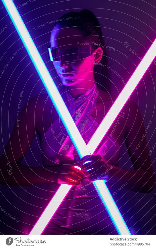 Futuristic young woman in trendy mirrored sunglasses with neon sticks in studio virtual reality futuristic self assured style confident portrait model