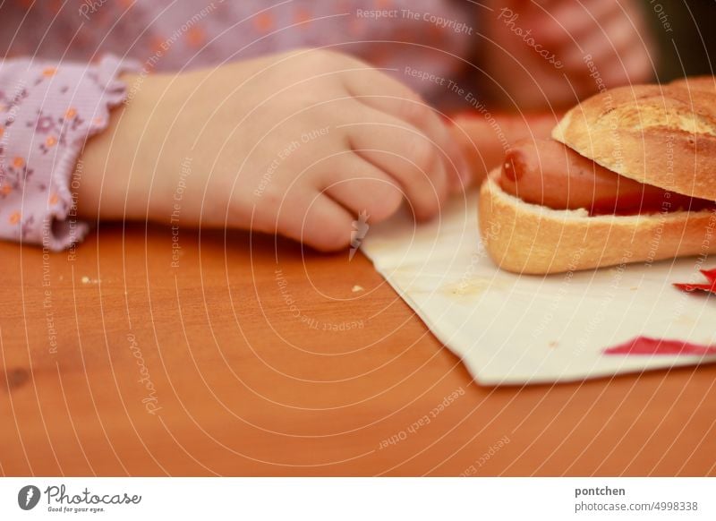 A hotdog lies on a table, beer table, in front of a child. Fast food Eating Child Hot dog Nutrition Food Unhealthy Snack bar Delicious Appetite Meal