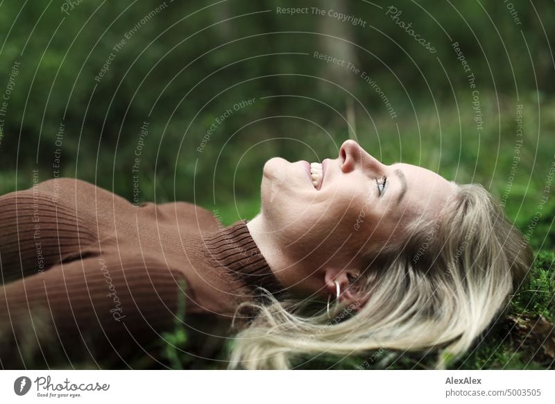 Young blonde woman with dimples lies on the moss in the forest and smiles at the camera from the side Young woman Woman Blonde Feminine pretty fortunate