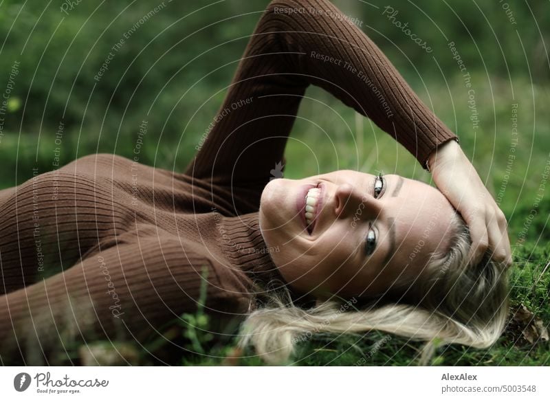 Young blonde woman with dimples lies on the moss in the forest and smiles at the camera from the side Young woman Woman Blonde Feminine pretty fortunate