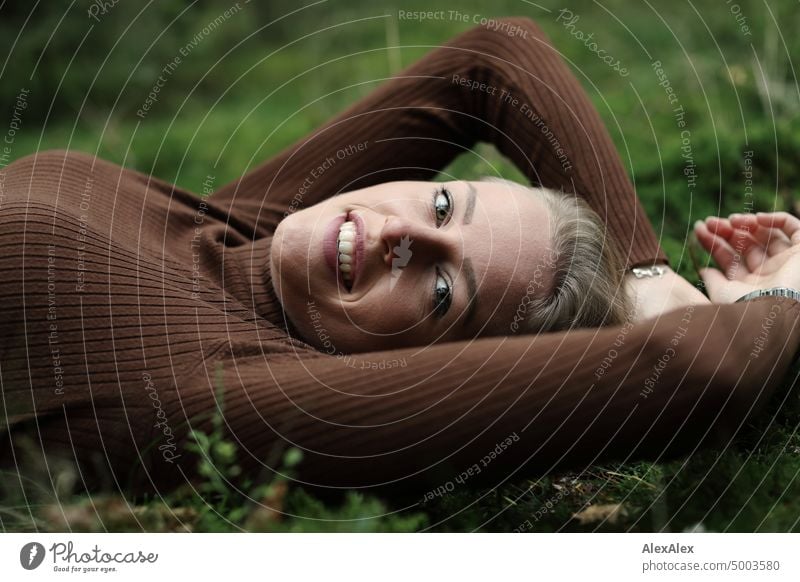 Young blonde woman with dimples lies on the moss in the forest and smiles at the camera from the side Young woman Woman Blonde Feminine pretty fortunate