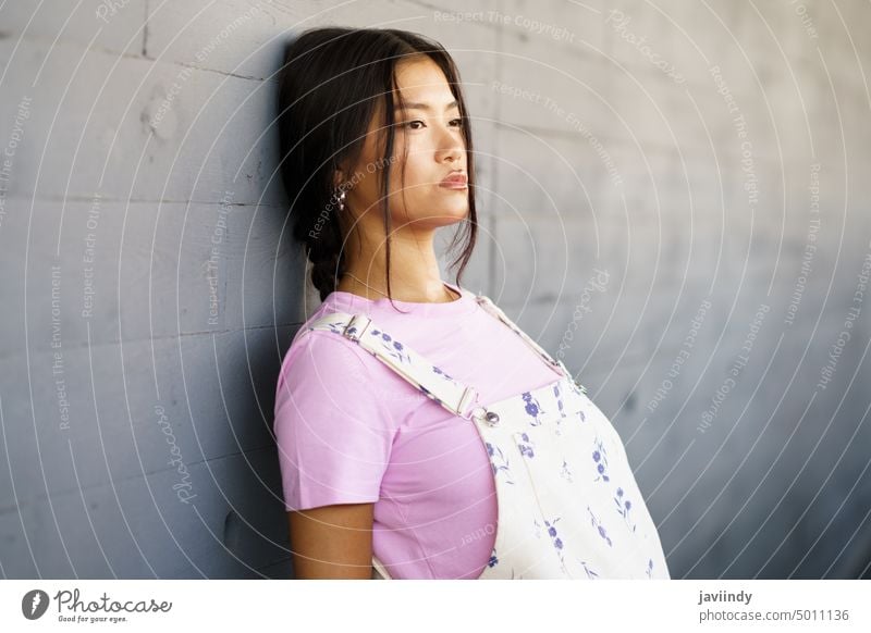 Young Chinese woman with a lost look and a serious expression leaning against the wall of a modern office building. asian chinese japanese fashion person girl