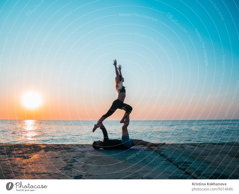 Fit young couple doing acro-yoga at sea beach. Man lying on concrete plates and balancing woman on his feet. Beautiful pair practicing yoga together. acroyoga