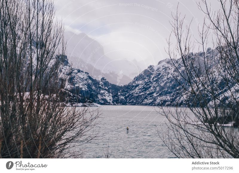 Man on paddle board between water and mountains on coast man floating tourist surface hill sup snow shore winter picturesque view stone nature travel landscape