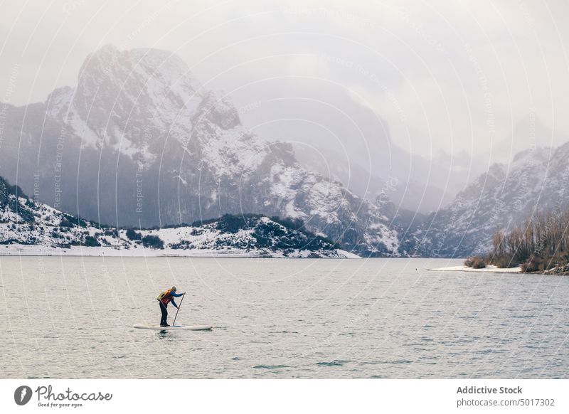 Man on paddle board between water and mountains on coast man floating tourist surface hill sup snow shore winter picturesque view stone nature travel landscape