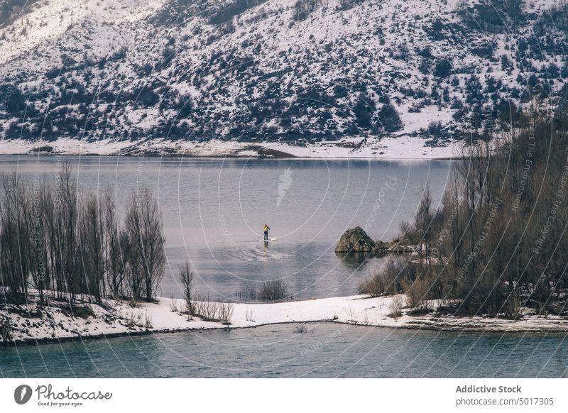 Man on paddle board between water and mountains on coast man floating tourist surface hill sup snow shore winter picturesque view stone nature travel landscape