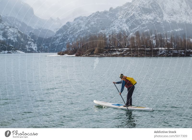 Man on paddle board between water and mountains on coast man floating tourist surface hill sup snow shore winter picturesque view stone nature travel landscape