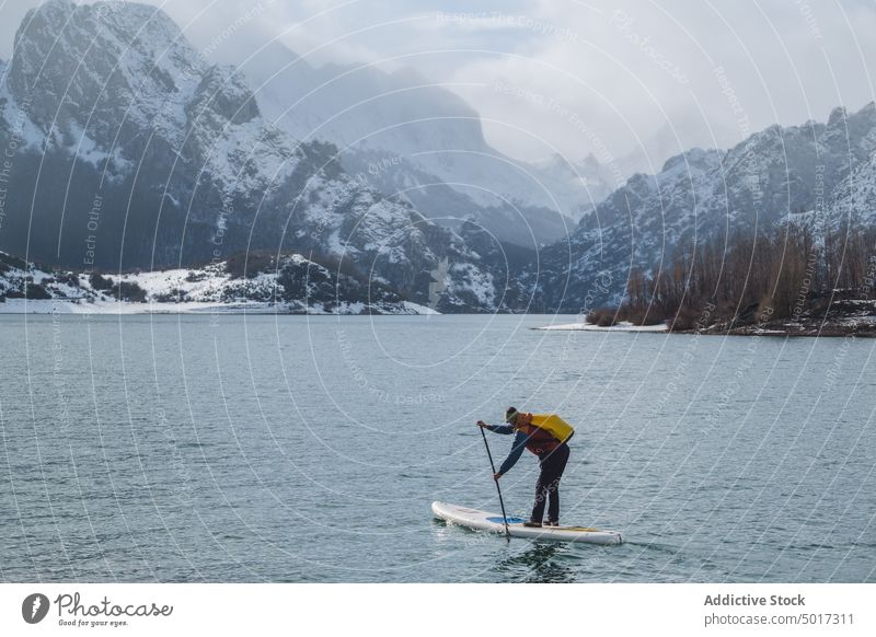 Man on paddle board between water and mountains on coast man floating tourist surface hill sup snow shore winter picturesque view stone nature travel landscape