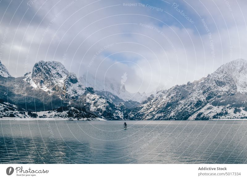 Man on paddle board between water and mountains on coast man floating tourist surface hill sup snow shore winter picturesque view stone nature travel landscape