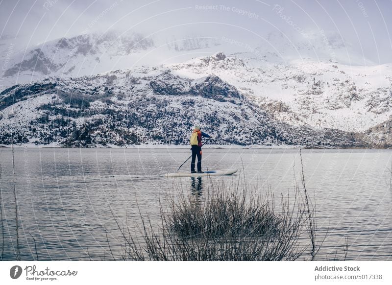 Man on paddle board between water and mountains on coast man floating tourist surface hill sup snow shore winter picturesque view stone nature travel landscape