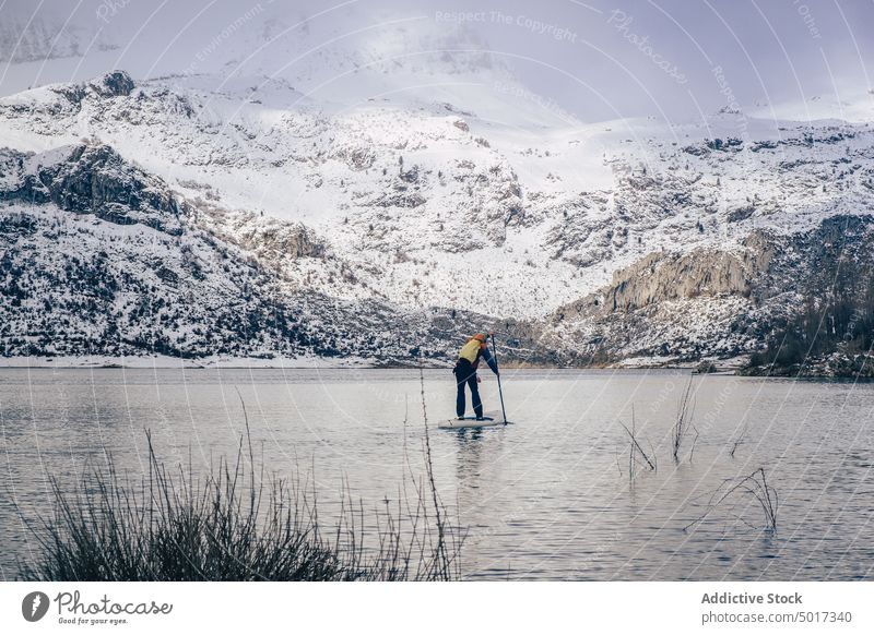 Man on paddle board between water and mountains on coast man floating tourist surface hill sup snow shore winter picturesque view stone nature travel landscape