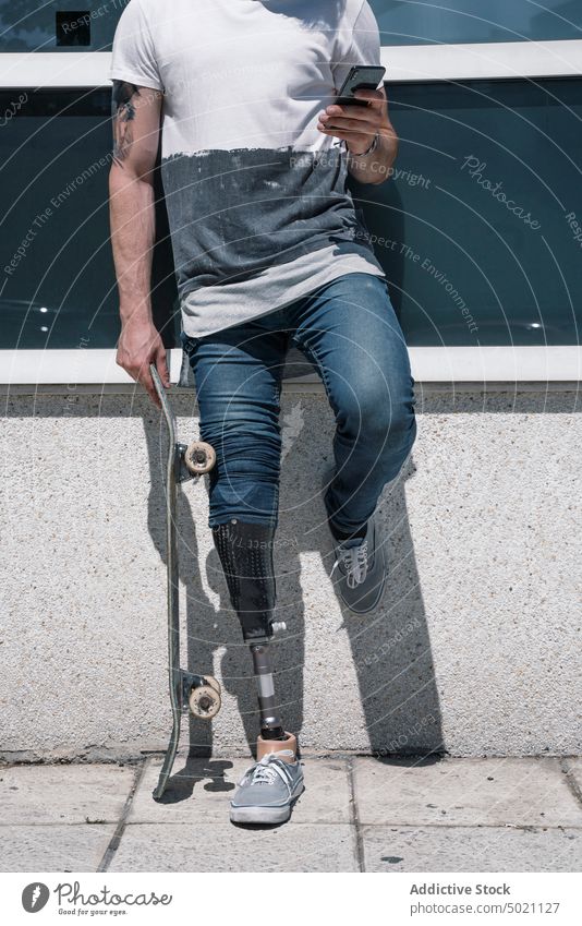 Crop skater with leg prosthesis using smartphone man skateboard artificial street city young urban male modern technology sunny daytime break social media hobby