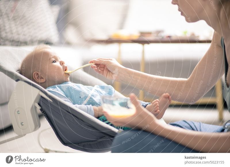 Mother spoon feeding her baby boy infant child in baby chair with fruit puree. Baby solid food introduction concept. healthy nutrition eating cute hungry dinner