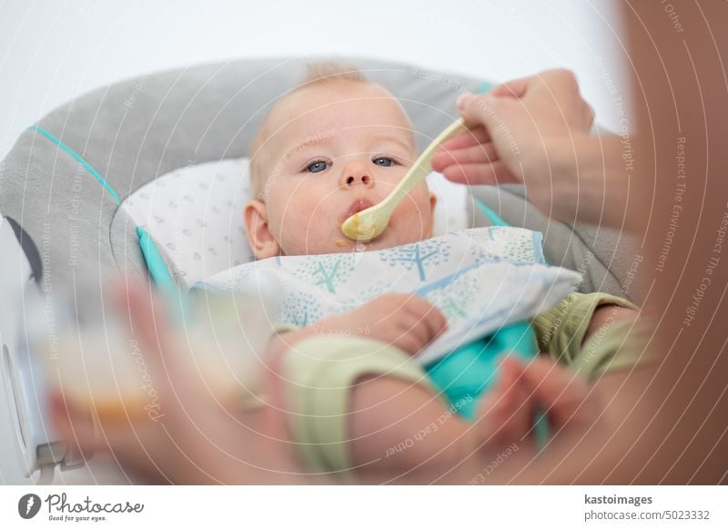 Mother spoon feeding her baby boy infant child in baby chair with fruit puree. Baby solid food introduction concept. healthy nutrition eating cute hungry dinner