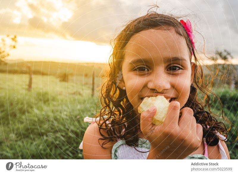 Hispanic girl eating apple in countryside nature rest sundown bite happy weekend summer child adorable field smile kid cute flower childhood joy curly hair