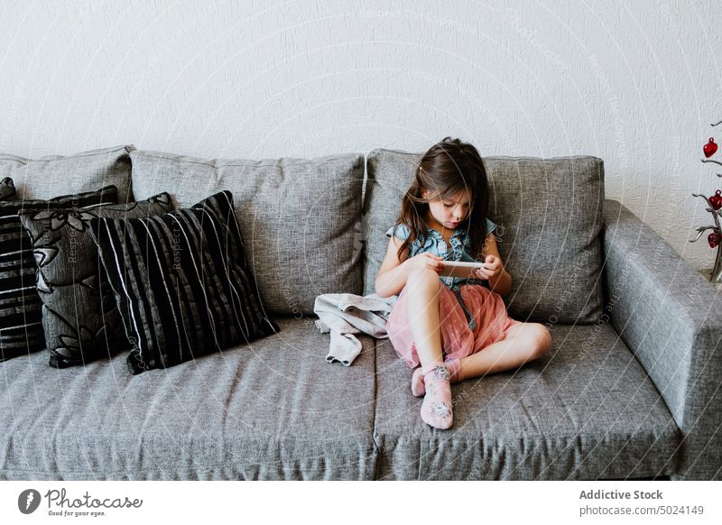 Cute ethnic child using smartphone on cozy couch at home play relax sofa comfort online internet videogame kid girl living room rest gadget casual dark hair