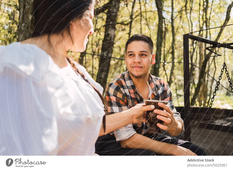 Ethnic woman passing mate to man in camp hot drink beverage infused interact calabash gourd spend time checkered shirt talk receive fresh bombilla speak content