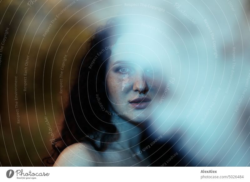 Young dark haired woman with curls and freckles looks half behind white steam cloud into camera in blue green light Woman Young woman Freckles Curl Brunette