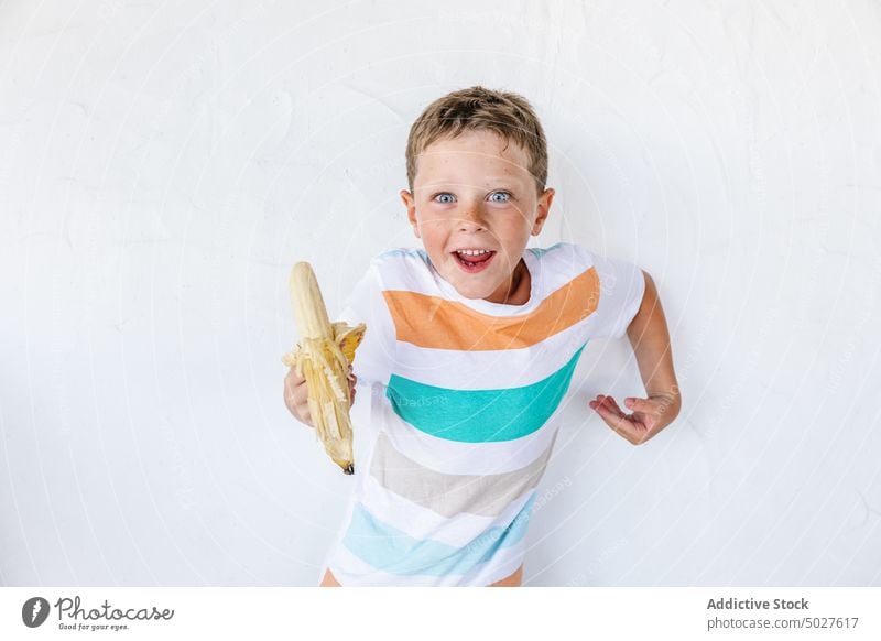 Joyful cute child eating banana and smiling leaning on white wall smile positive fruit boy happy vitamin adorable portrait fresh childhood glad kid joy t shirt