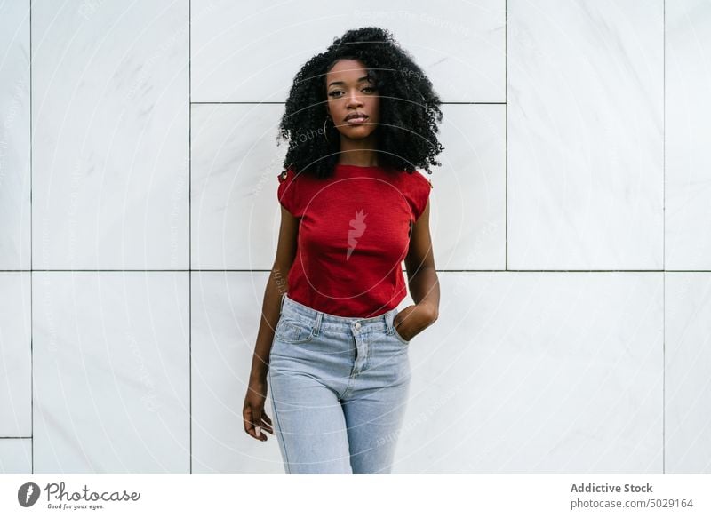 Unemotional ethnic woman standing near tiled wall unemotional serious building hand in pocket makeup appearance afro casual style outfit female african american