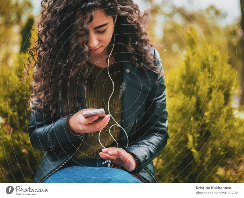 Pretty young latin girl with black curly long hair listening music with earphones and smartphone or player on a deserted autumn street. Woman enjoying moment.