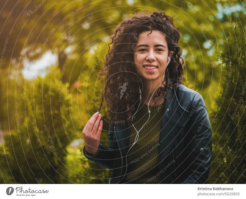 Pretty young latin girl with black curly long hair listening music with earphones and smartphone or player on a deserted autumn street. Woman enjoying moment.