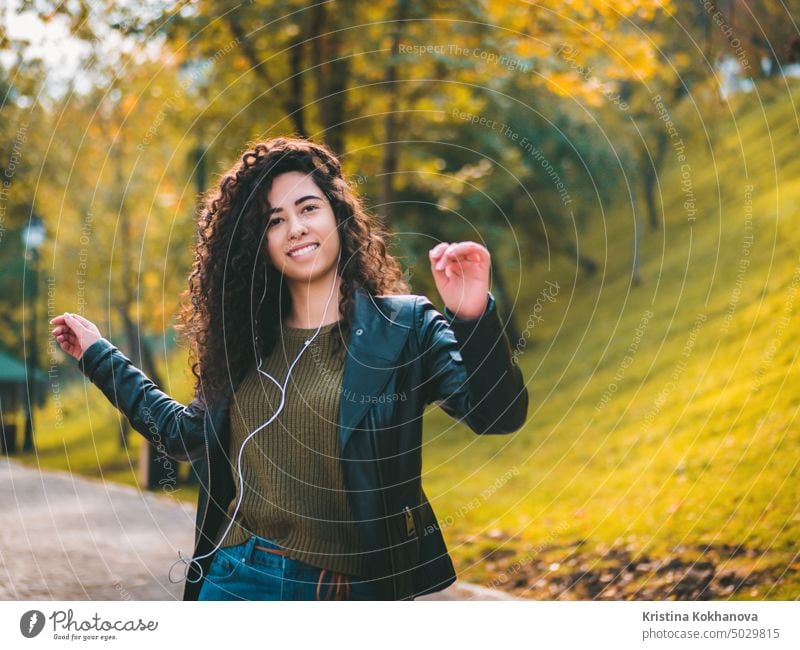 Happy african young woman with curly hair listening to music on earphones. Hispanic hipster girl dancing to rhythm and singing along melody in the autumn park.