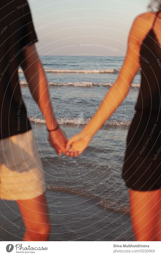 Unrecognizable couple holding hands on the beach. Man and woman together with waves of sea on background. Closeup framing. Tanned skin at sunset. Love and relationships. Summer love story of young adults. Millennial on vacation.