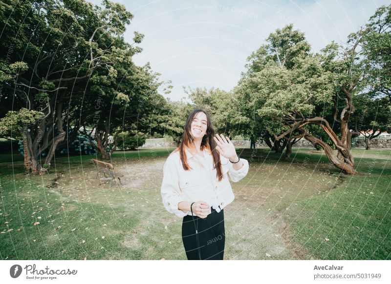 Portrait young woman greeting to camera during a sunny day at the park, wide angle lens image person smile stylish youth female happy carefree elegant enjoy