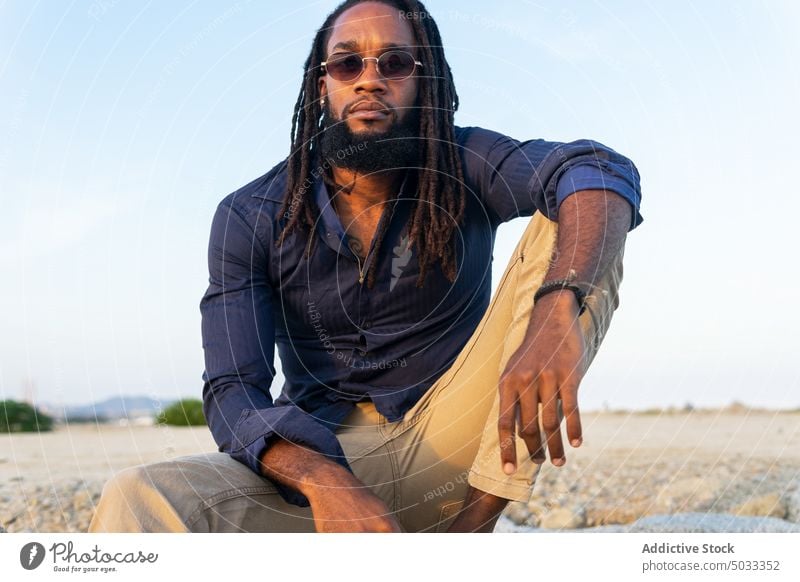 Stylish black male sitting on beach in sunshine man coast chill rest style shore individuality personality portrait dreadlocks cool hairstyle sunglasses guy