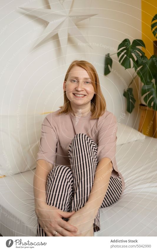 Smiling woman in striped pants sitting on bed positive smile bedroom comfort lamp monstera star at home female domestic young happy light apartment lady green