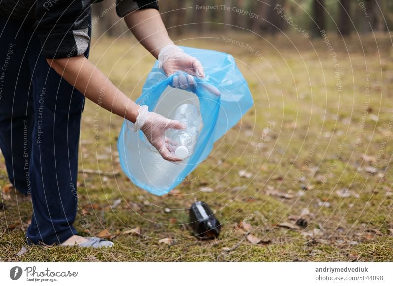 A woman hands in gloves collects and puts used plastic bottle into a blue trash bag. A volunteer cleans up the park on a sunny bright day. Clearing, pollution, ecology and plastic concept