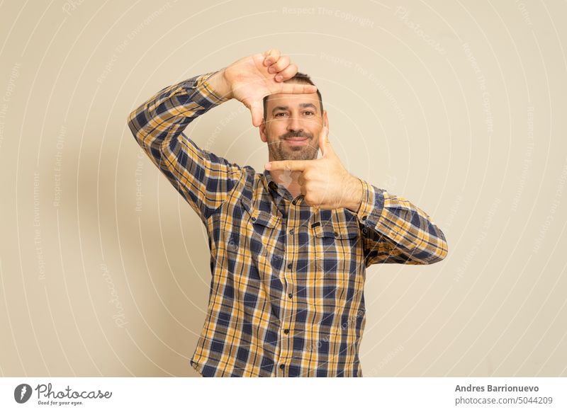 Handsome bearded happy male blinks his eyes and looks through a frame formed by his hands. An attractive man makes a frame with his fingers, isolated on a beige background with copy space for text. people concept.