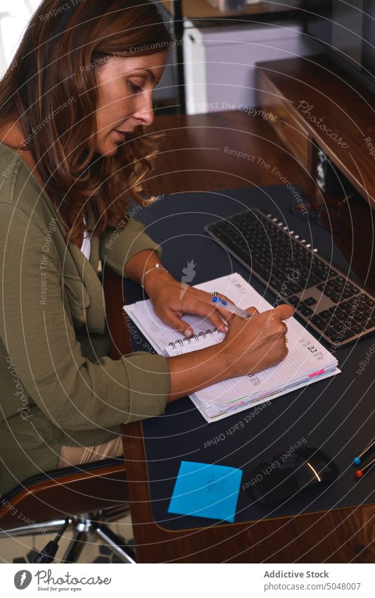 Focused lady writing in notepad in office woman take note write freelance computer work self employed independent notebook table female pastime fair hair casual