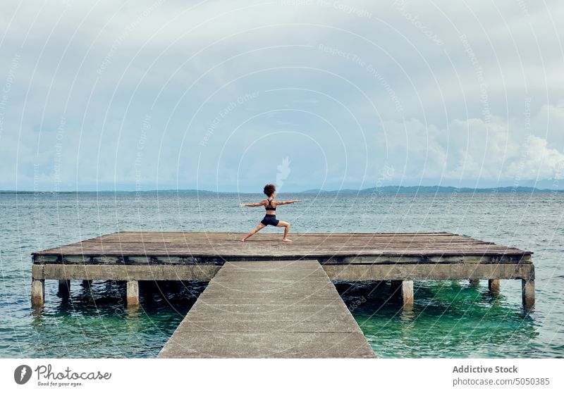 Woman doing yoga on shabby pier in sea woman meditate zen asana virabhadrasana warrior pose balance peaceful wellness quay tranquil recreation mindfulness