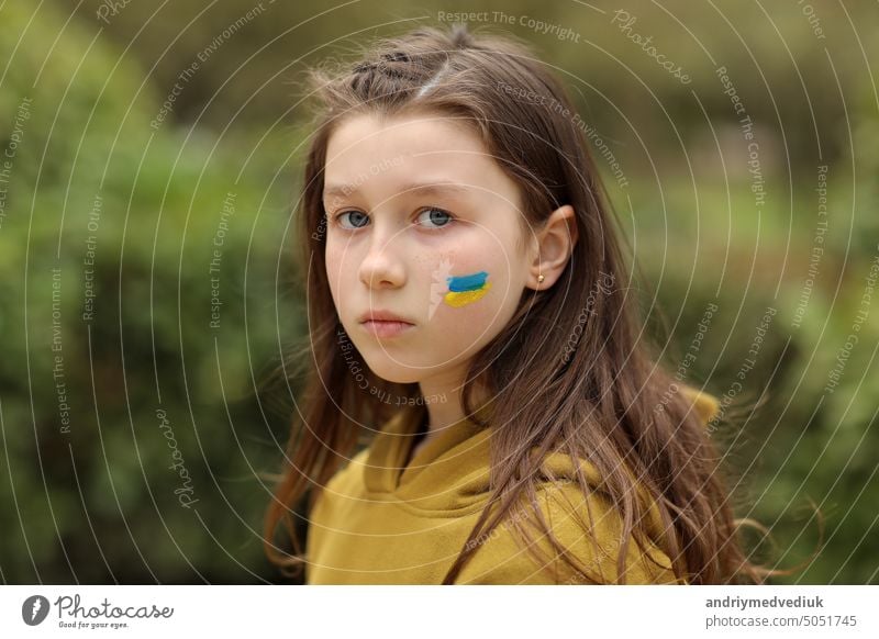 the face of a frightened girl, painted on her cheek in the yellow-blue colors of the Ukrainian flag, a request for help. Children ask for peace. High quality photo. High quality photo