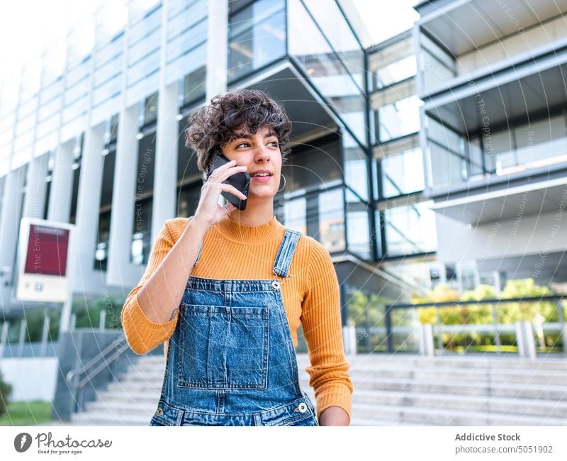 Satisfied female on a phone call against building on sunny day woman smartphone using audio talk speak street summer urban style city smile positive brunette