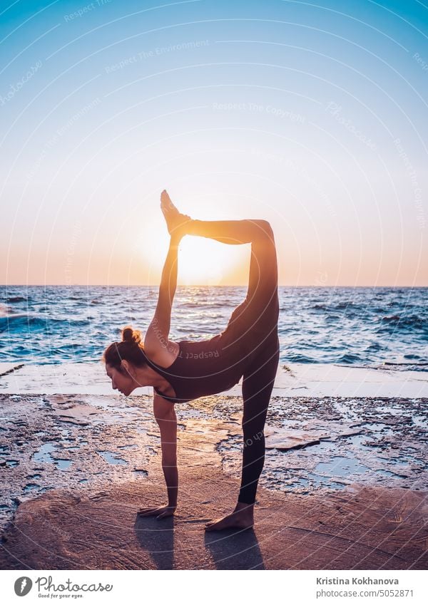 Slim woman in black bodysuit practicing yoga near sea or ocean during sunrise light. Flexibility, stretching, fitness, healthy lifestyle. abstract active adult