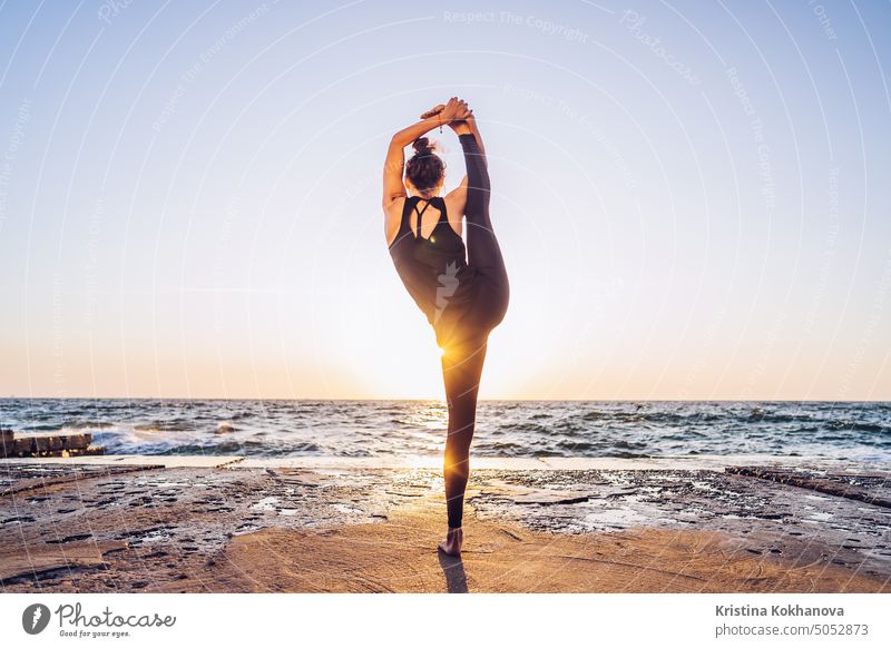 Slim woman in black bodysuit practicing yoga near sea or ocean during sunrise light. Flexibility, stretching, fitness, healthy lifestyle. abstract active adult