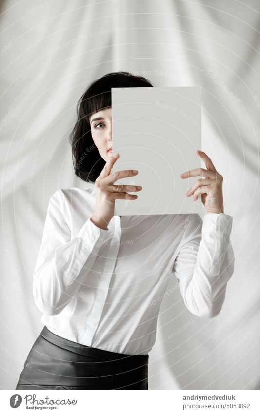 beautiful young businesswoman with dark short hair holding a magazine, book with mock up. girl in a white shirt and black leather skirt standing in studio